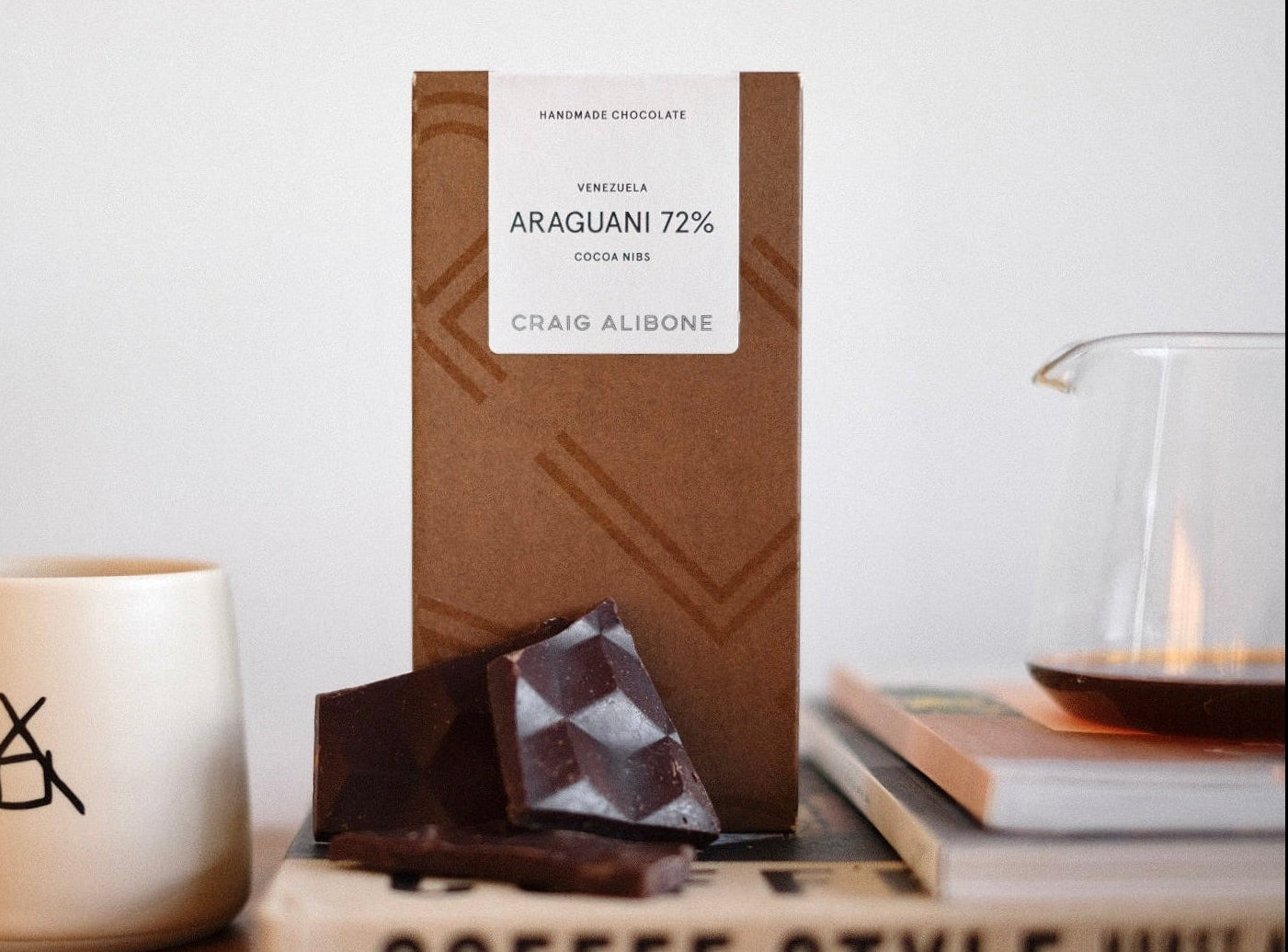 Chocolate bar with a label on a white surface, next to a mug and books.