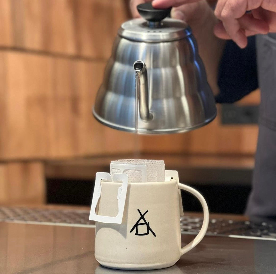 Person pouring coffee from a silver kettle into a white mug with a  the barn drip bag inside on a wooden surface.