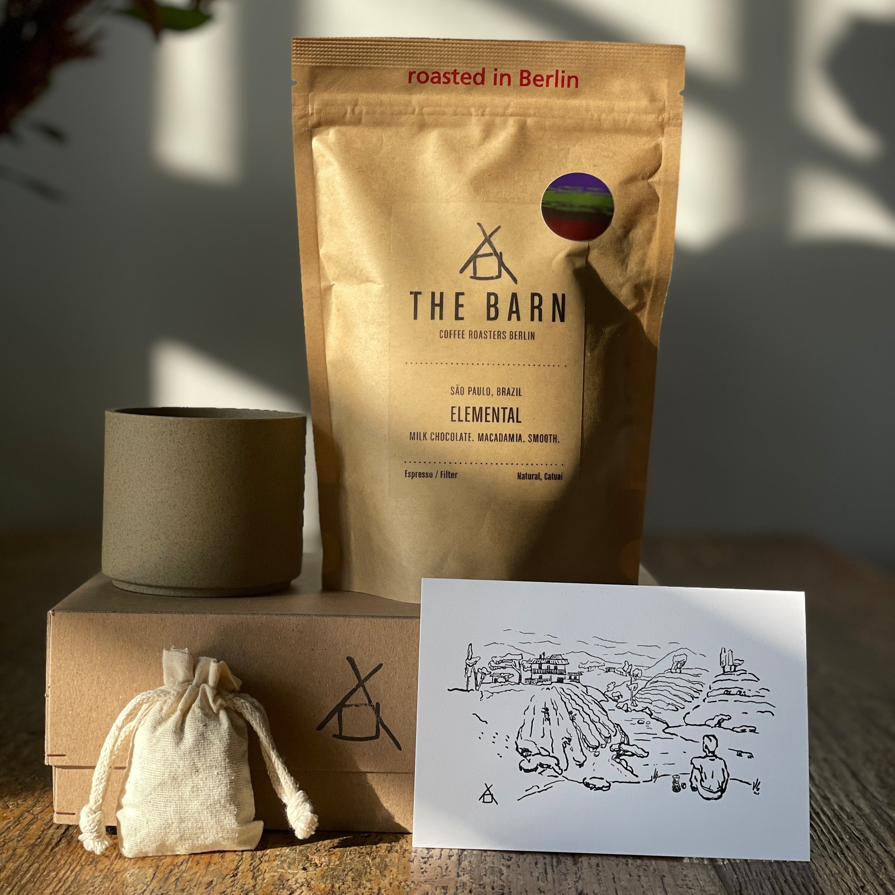 A gift box setup with a bag of coffee beans labeled 'The Barn', a beige Hasami mug, and a small cloth drawstring bag, all placed on a wooden surface with a printed black and white illustration next to them.