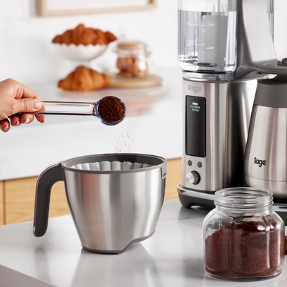 Person measuring coffee grounds into a Sage Luxe Brewer™ Thermal Coffee Maker on a kitchen counter.