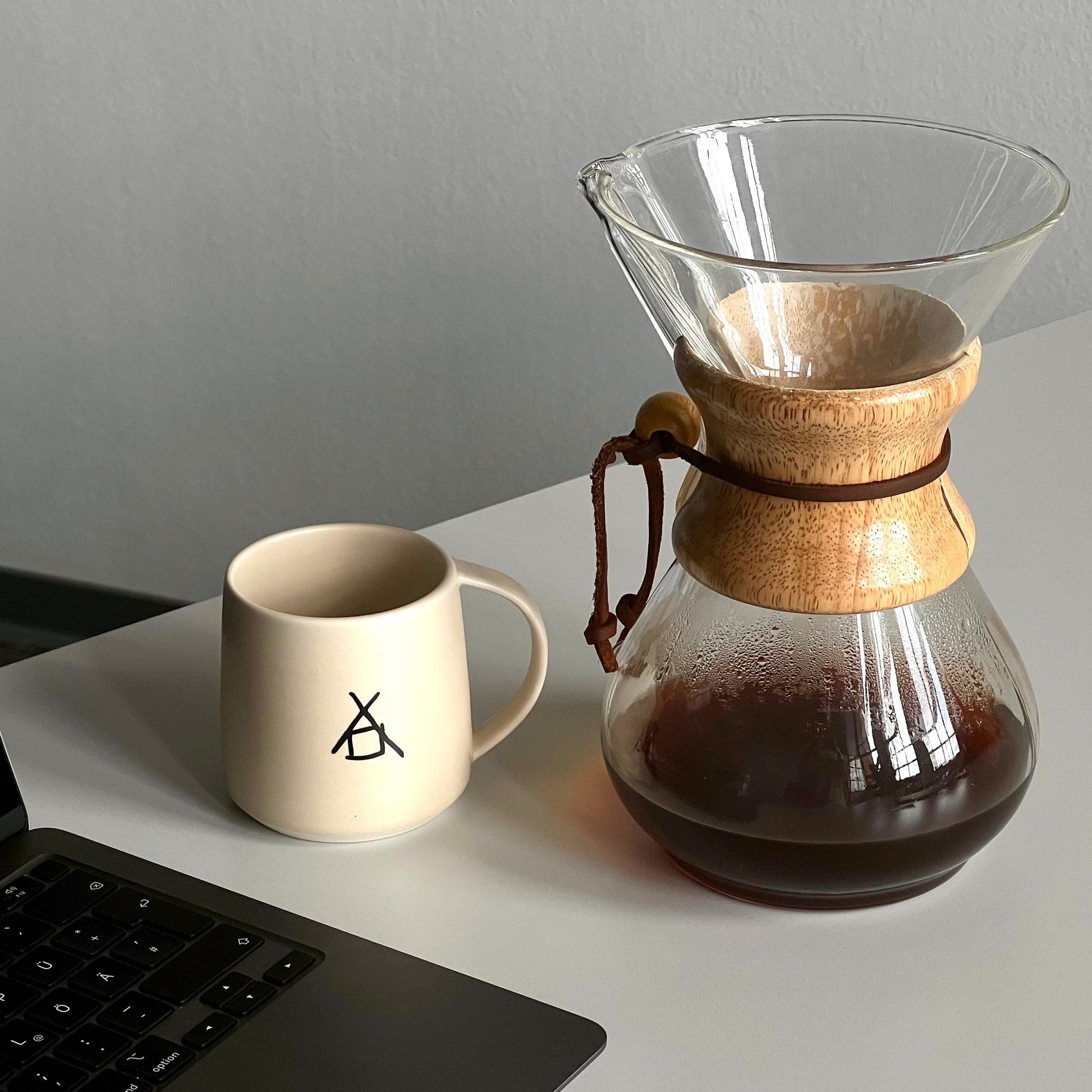 Ein offener Laptop steht außerhalb des Bildes, daneben ein weißer Becher mit dem The Barn-Logo. Neben dem Becher steht eine Chemex-Kaffeemaschine, die zur Hälfte mit frisch gebrühtem Kaffee gefüllt ist.