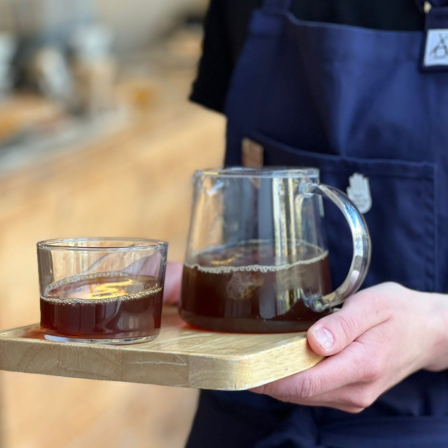 A person's hand is holding a wooden tray with a glass cup and glass server filled with freshly roasted specialty coffee