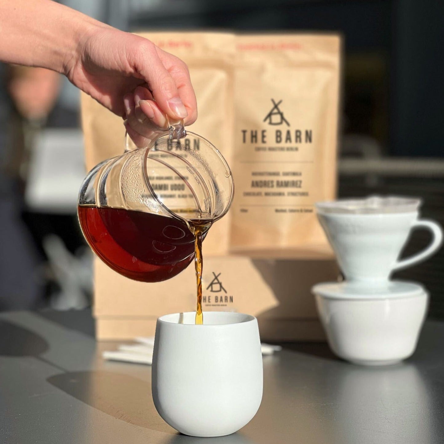 A person pouring freshly roasted coffee from a glass server into a ceramic mug with Hario dripper set and THE BARN curated subscription in the background