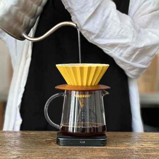 Person pouring water from a stainless steel kettle into a Yellow Origami Dripper with a brown wooden holder over a glass server